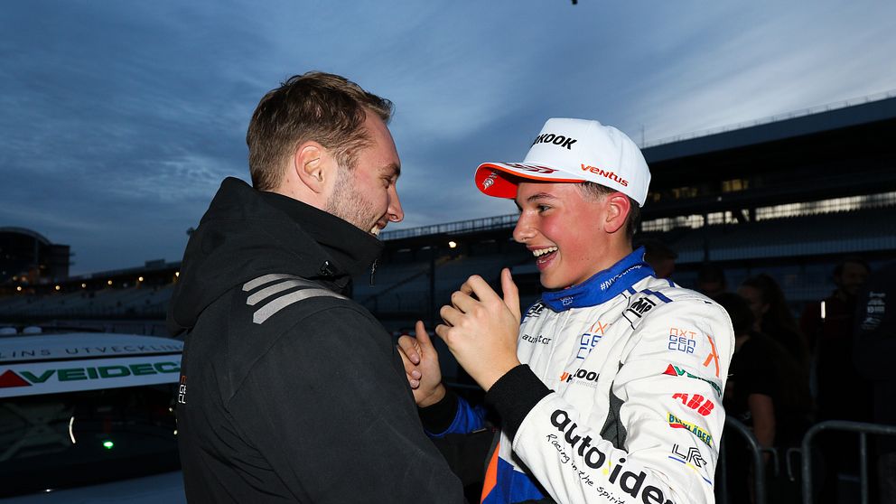 Två personer interagerar i en pitområde vid en racinghändelse. Den ena personen i en mörk jacka står nära en annan person iförd en motorsportoverall med sponsorlogotyper. Bakgrunden visar en racingbil och en dimmig himmel som antyder skymning.