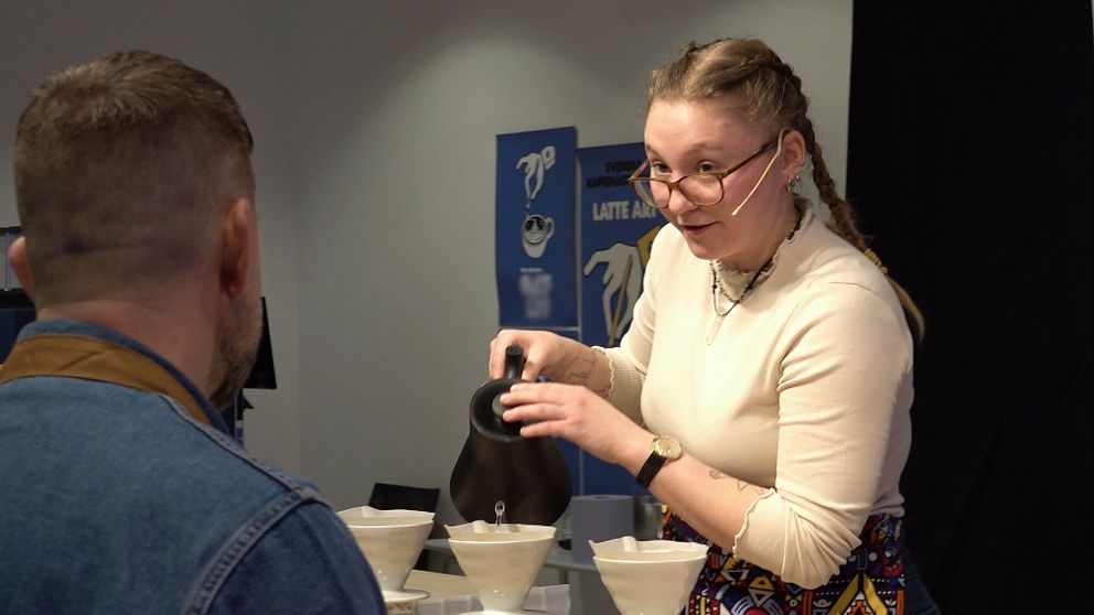 En kvinna häller kaffe från en kanna i vita filter på ett bord medan en man med jeansjacka tittar på, i vad som verkar vara ett kaffeevenemang eller en kaffedemonstration.