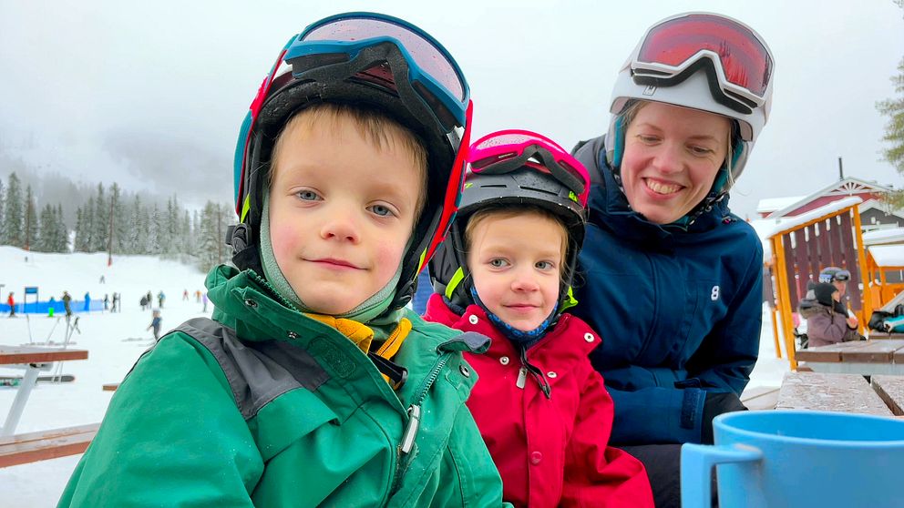 Två yngre barn och en mamma i skidkläder och hjälmar frmför en skidbacke