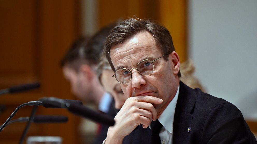 Statsminister Ulf Kristersson i kostym lyssnar under ett möte med mikrofoner och suddiga personer i bakgrunden.