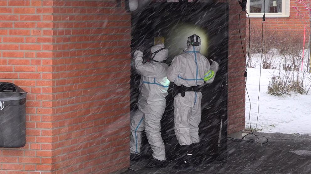 Två personer – poliser – klädda i skyddsutrustning arbetar vid en ingång till en byggnad, ett soprum, i snöigt väder. En av dem håller upp en lampa, vilket skapar en ljuspunkt i det mörka utrymmet inuti. I bakgrunden syns en soptunna och snö på marken.