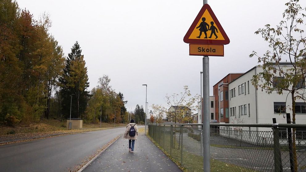 En flicka går på en gata, bredvid en skylt där det står ”skola”