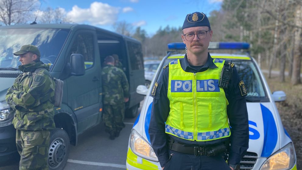 Bild på Polis som står framför en polisbil med hemvärnssoldater i bakgrunden.