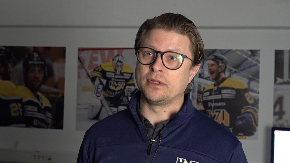 Hockeytränare med blå jacka framför tre matchfotografier på väggen.