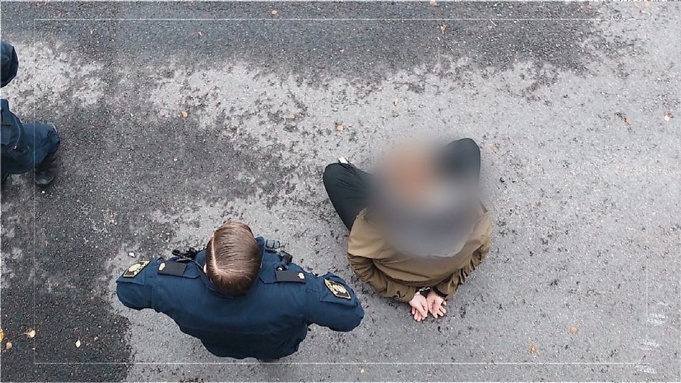 En person sitter på marken med händerna handfängslade bakom ryggen. Bredvid står en man i polisuniform.
