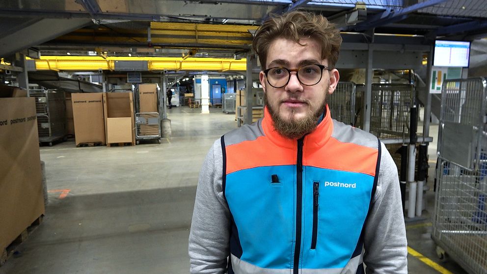 En person står på en logistik- eller distributionsanläggning, iförd en färgglad arbetsjacka med ett tryck av ”Postnord”. I bakgrunden syns pallar och lagerutrustning, vilket tyder på en intensiv arbetsmiljö för post- och paketdistribution. Belysningen är stark och det finns flera sektioner av lagringsutrymme.
