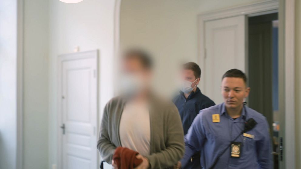En person går in i ett rum, följd av två säkerhetsvakter. Rummet har ljusa väggar och en dörr med en enkel design i bakgrunden. Den ena säkerhetsvakten är klädd i en blå uniform och har en namnskylt, medan den andra bär en grå tröja. Atmosfären i bilden antyder en offentlig miljö, eventuellt en myndighet eller en institution.