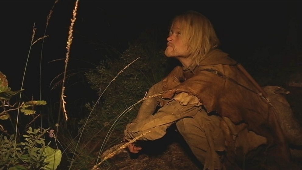 Person i bruna kläder hukar i en mörk skogsmiljö.
