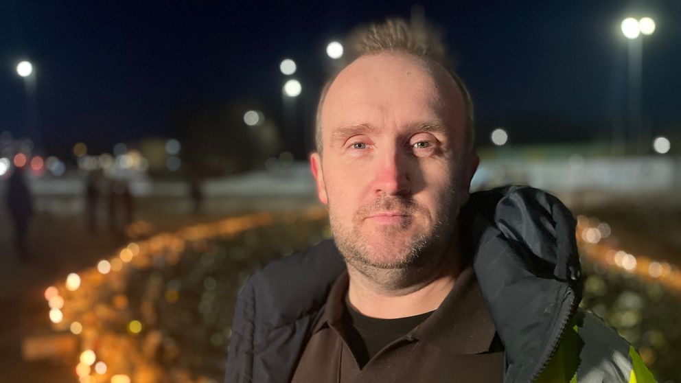 En person iklädd en reflexjacka står i förgrunden. I bakgrunden syns ett ljushav av tända ljus vid en minnesplats utomhus i kvällsmörkret.