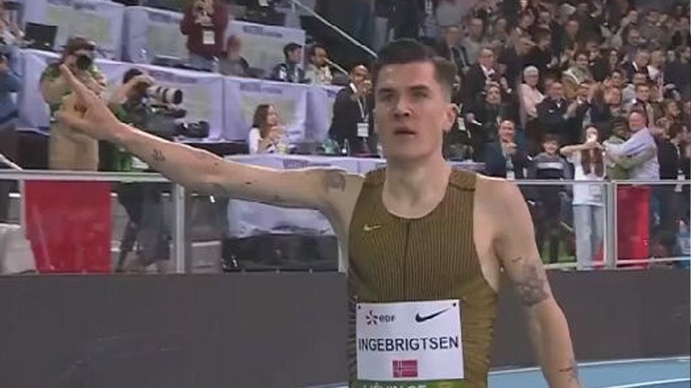 En idrottare iförd en bronsfärgad tävlingsdräkt från Nike står på en inomhusbana, med publiken i bakgrunden och sträcker ut ena armen.