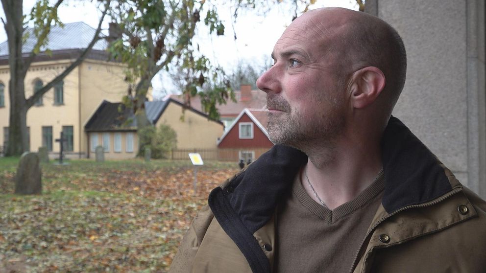 Man i beige jacka står utomhus vid en kyrkogård med gula och röda byggnader i bakgrunden.
