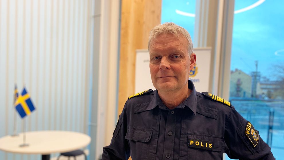 En polisman tittar in i kameran och håller handen på ett bord. Bakom honom syns två Sverigeflaggor.