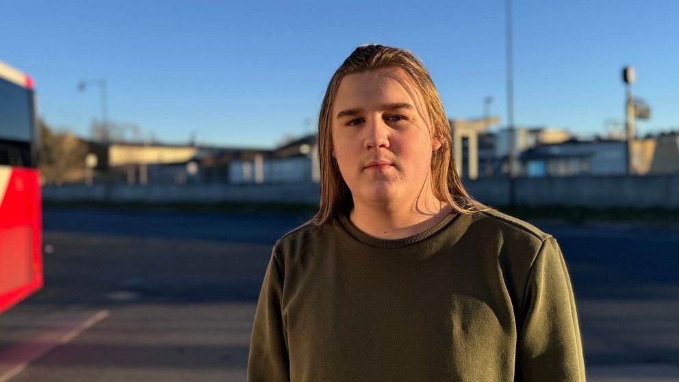En person står framför en buss på ett resecentrum, omgiven av en klarblå himmel och tågrälsen i bakgrunden. Personen är klädd i en grön tröja och blicken är riktad mot kameran. Bussens färgschema inkluderar röd och vit.