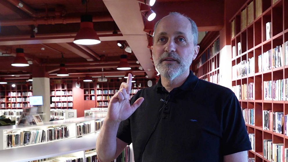 Man, Lars Alkner, står i ett bibliotek och gör en pistolgest med handen.