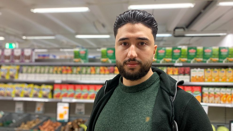 En man står i en matbutik och tittar rakt in i kameran. Grönsaker och andra varor i bakgrunden.