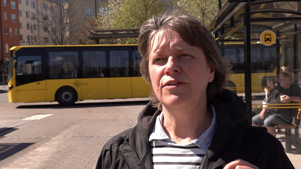 En kvinna står vid en busshållplats och en gul buss syns i bakgrunden.