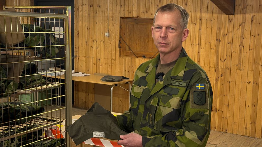 man i militäruniform håller fram en grön BH i ett förråd med träväggar