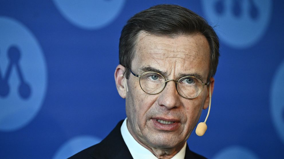Statsminister Ulf Kristersson i kostym framför blå bakgrund med Moderaternas logo.