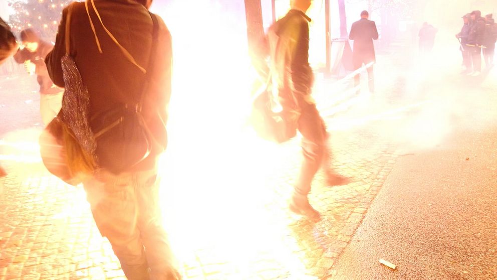 Personer som skyndar sig förbi en explosion eller kraftig ljuseffekt i en stadsmiljö, med rök och ljus som sprider sig i luften. Många verkar vara på väg bort från händelsen, vilket skapar en känsla av hastighet och oro.