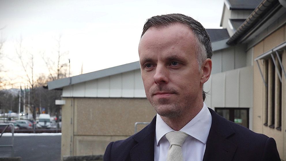 Åklagare David Kahl står utomhus klädd i kostym med en vit skjorta och slips, med en byggnad i bakgrunden och träd utan löv i förgrunden som indikerar tidig vinter.
