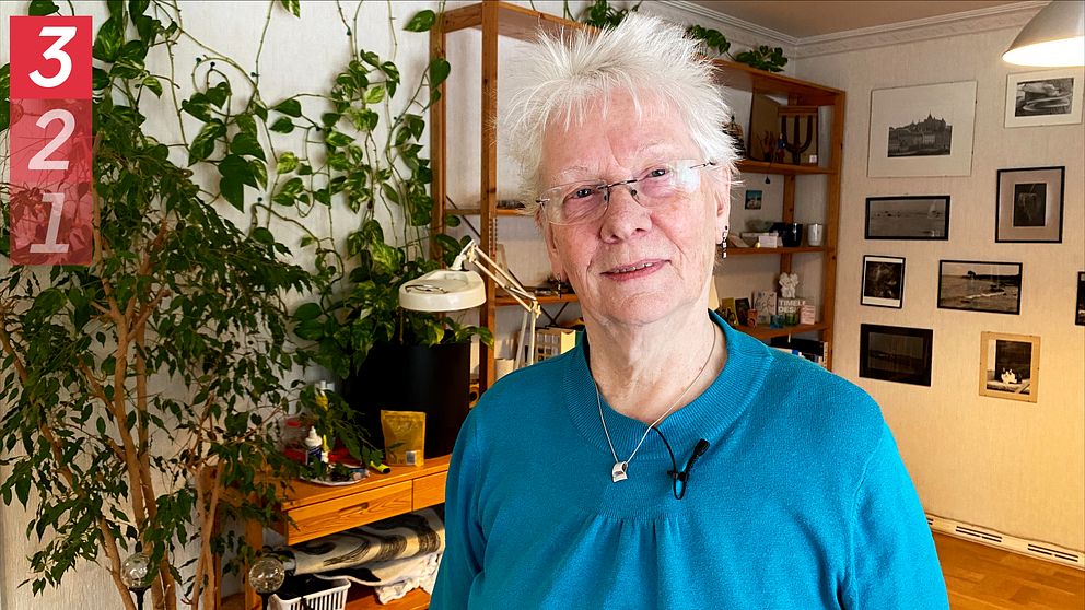 Anita Kullander, en psykolog, står i sitt vardagsrum med bokhylla och hängväxt bakom på väggen.