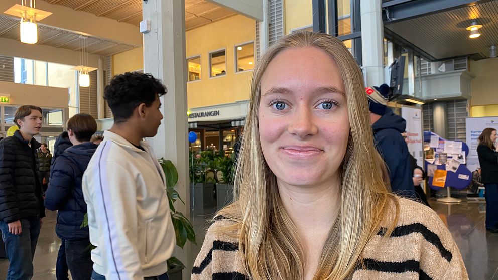 En person i en randig tröja står inomhus på Mittuniversitetet och det är liv och rörelse i omgivningen.