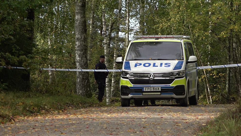 En polisbil står på en grusväg omgiven av träd, med avspärrningsband som markerar en plats för en pågående utredning. En polis fungerar som vakt vid avspärrningen. Höstlöv ligger på marken.
