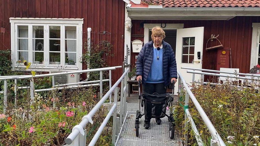 Solveig Ryd som är missnöjd med Blekinges nya trygghetslarm står med sin rollator utanför sitt röda hus med vita knutar.