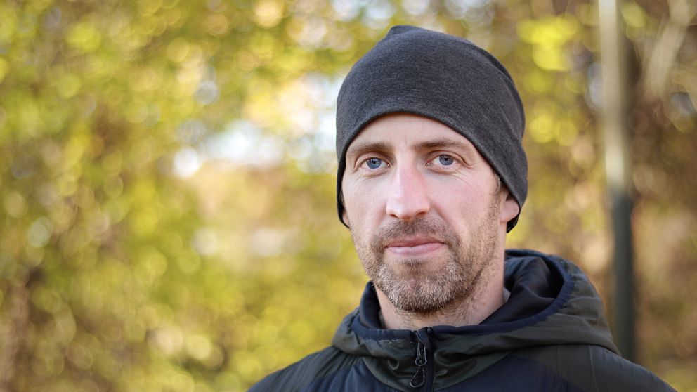 En person står i en grönskande utomhusmiljö, klädd i en svart jacka och en mörk mössa. Bakgrunden är oskarp men visar träd och höstfärger, vilket skapar en mjuk och naturlig atmosfär.