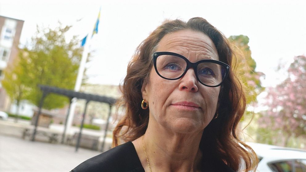 Kvinna med långt brunt hår står utomhus, svensk flagga syns i bakgrunden.