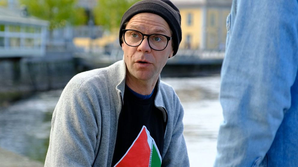 Person med grå kofta och svart t-shirt sitter utomhus vid vattnet, samtalar med annan person.