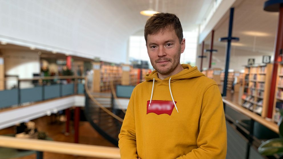 En man tittar rakt in i kameran. Han har en gul tröja och i bakgrunden är det ett bibliotek.