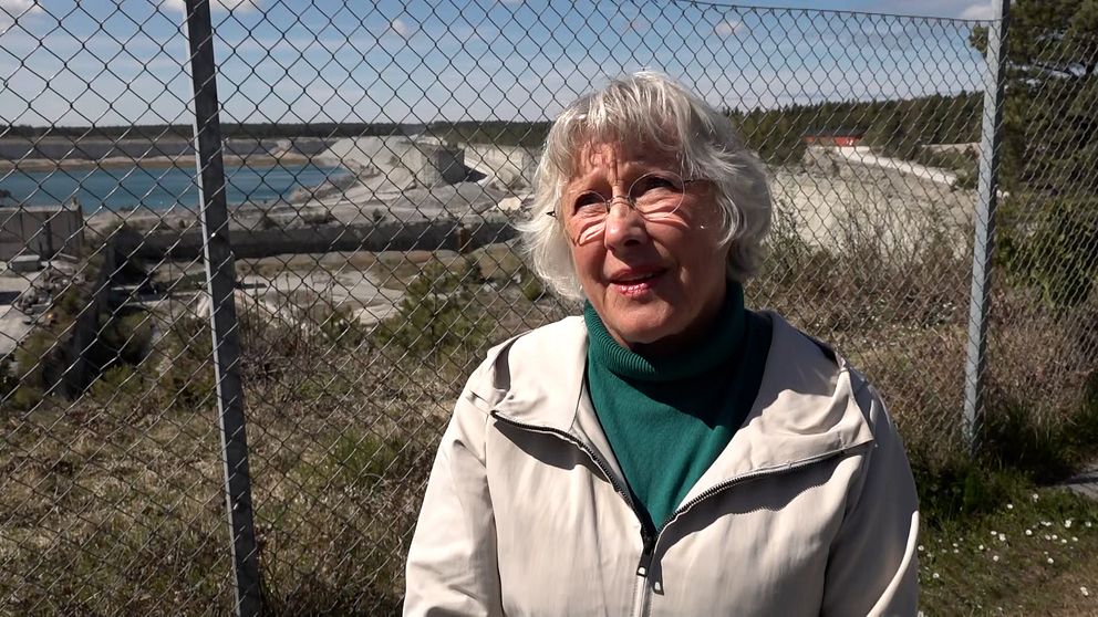 Person med vitt hår står utomhus vid stängsel, hav och industriområde syns i bakgrunden.