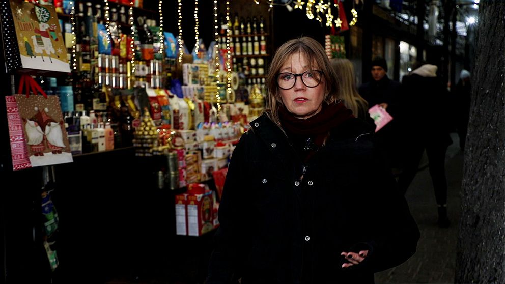 Person står framför en färgglad julmarknad med olika presenter och dekorationer i bakgrunden, belyst av julbelysning.