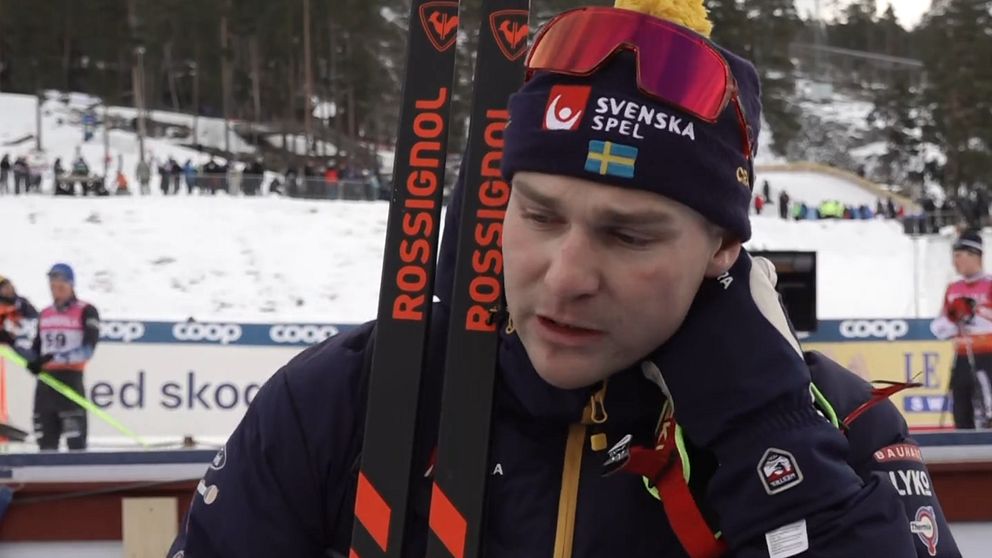 En skidåkare i tävlingsdräkt håller i sina skidor med Rossignol-logotyp. I bakgrunden syns en vintersportarena med snö och andra åkare.
