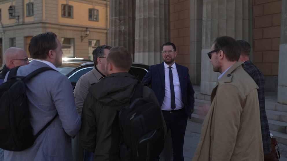 En grupp män i samtal utanför en byggnad med höga pelare i stadsmiljö.