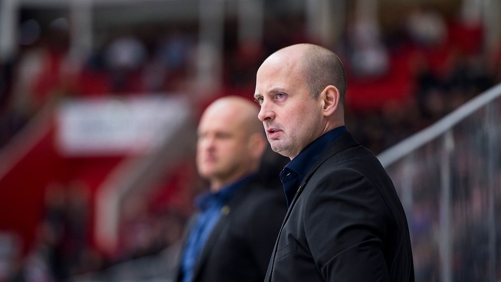 Två män står vid sidan av en hockeyrink, klädda i kostymer. I bakgrunden syns en publik i röda läktarsektioner, vilket skapar en sportig atmosfär. Den ena mannen ser ut att vara i fokus, medan den andra står lite längre tillbaka.