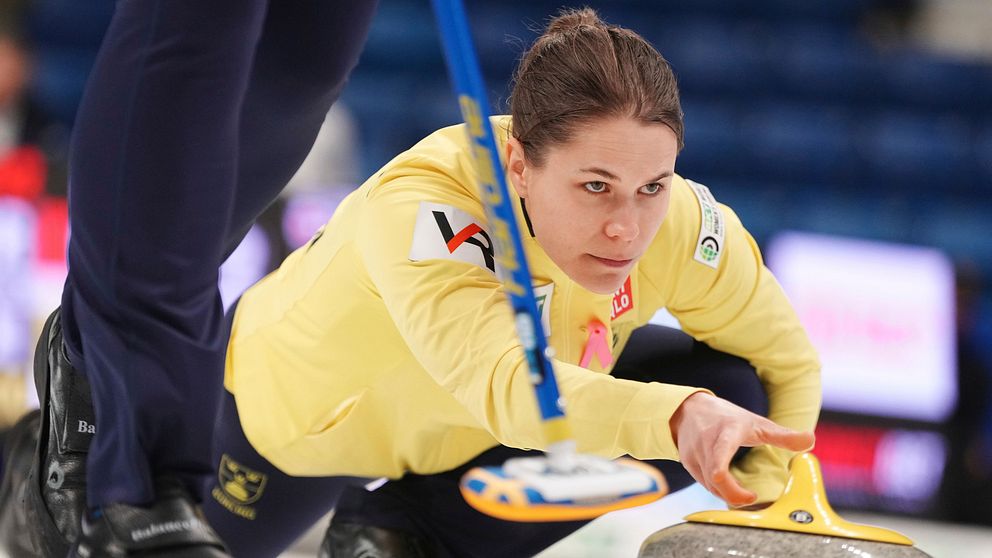 Anna Hasselborg i en gul tröja ligger på knä och siktar på en curlingsten som glider över isen. En annan spelare syns delvis i bakgrunden, klädd i mörka byxor. Hallen är inredd för en curlingmatch med oskarpa åskådare i bakgrunden.