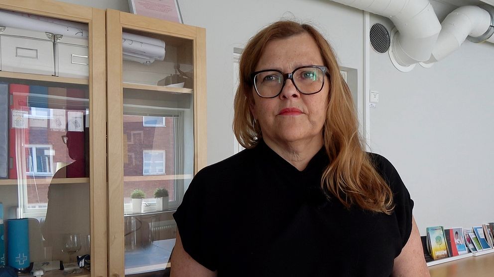 En kvinna med långt brunt utslaget hår som faller över vänster axel, hon har svarta glasögon och svart v-ringad tröja som går högt upp mot halsen.