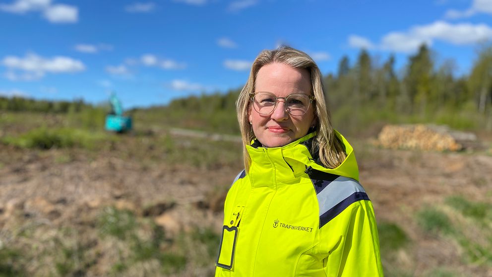 Person i gul varseljacka står utomhus på kalhygge, grävmaskin syns i bakgrunden.