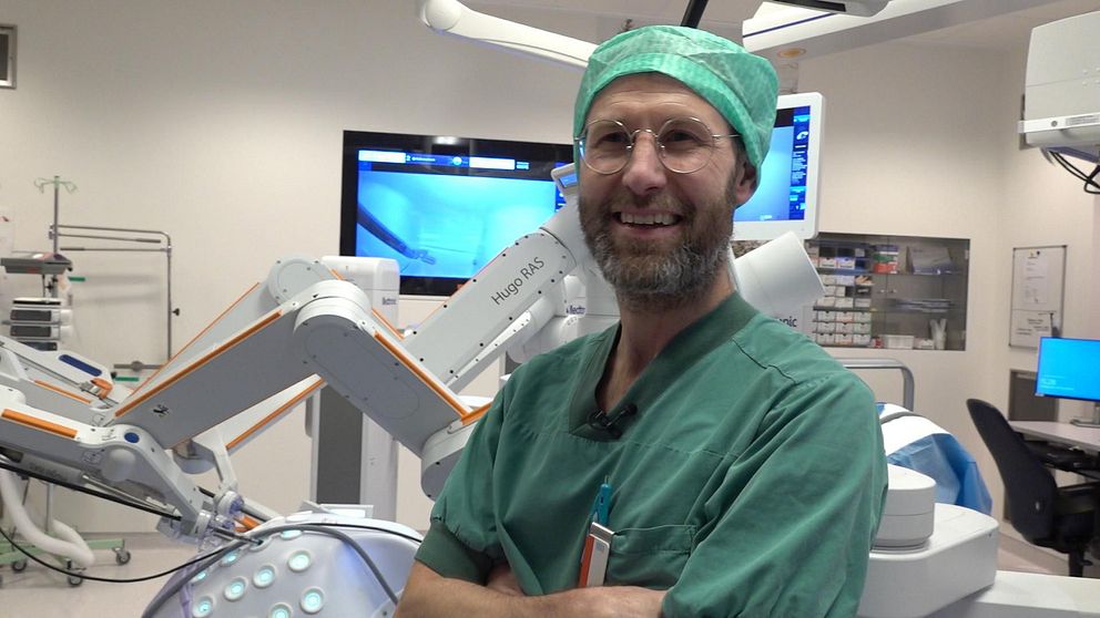 Överläkaren Mattias Tell står leende i gröna operationskläder framför en maskin – en operationsrobot