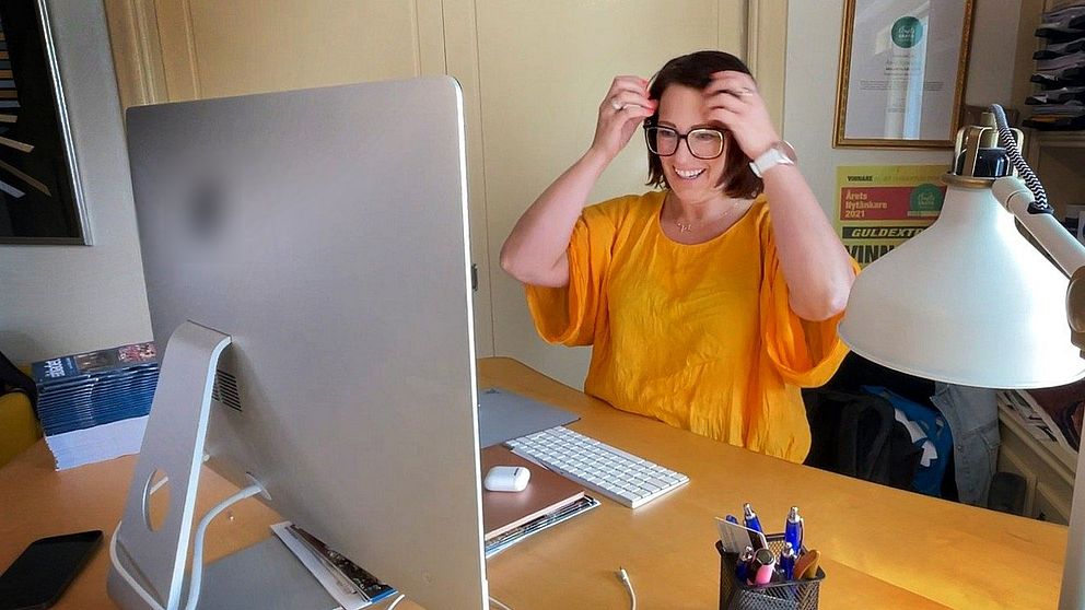 Person i gul tröja sitter vid skrivbord med dator, lyfter upp en surfplatta eller bok.