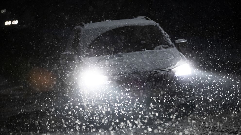 En bil kör med tända framljus i mörkret. Omkring bilen faller snön.