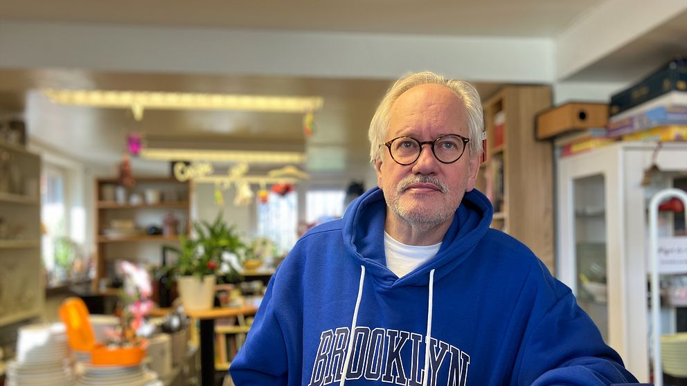 En äldre man i en blå Brooklyn-hoodie står i en återbrukslokal med uppställda tallrikar och inredning i bakgrunden.