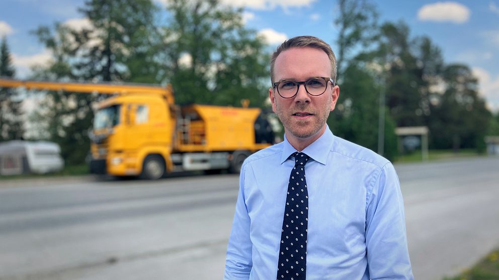 Andreas Carlsson, kd, infrastrukturminister, står framför en väg och framför ett fordon som kan laga vägar.
