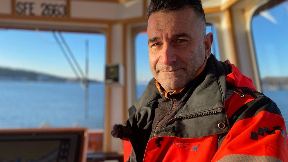 Man i orangea yrkeskläder står i fören på en båt och havet syns bakom honom.
