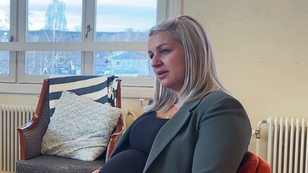 En gravid kvinna sitter på en stol med en grön kavaj och en svart topp, och hon ser ut över ett rum med fönster som ger utsikt mot en stadsmiljö. I bakgrunden finns en soffa med mönstrat tyg och en radiator. Rummet har en ljus och neutral inredning.