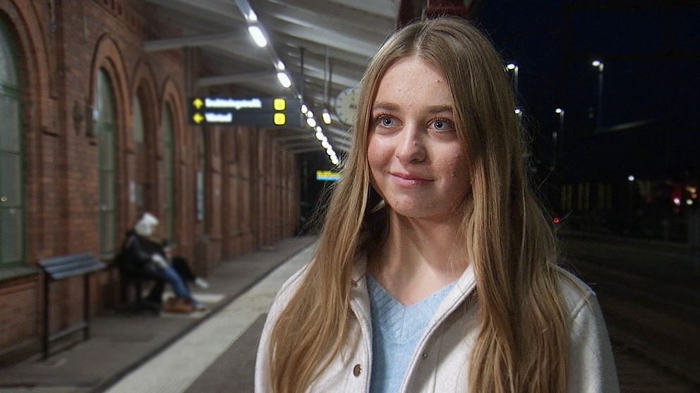 En ung kvinna med långt hår står på en tågstation. I bakgrunden syns ett bänkar och en passagerare som väntar.