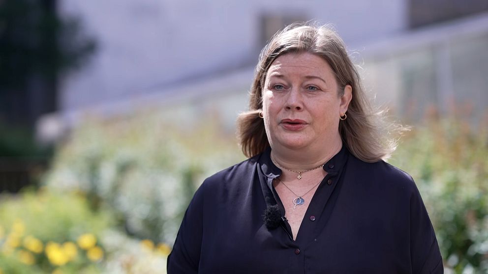 Person med långt blont hår står utomhus framför en grönskande bakgrund.