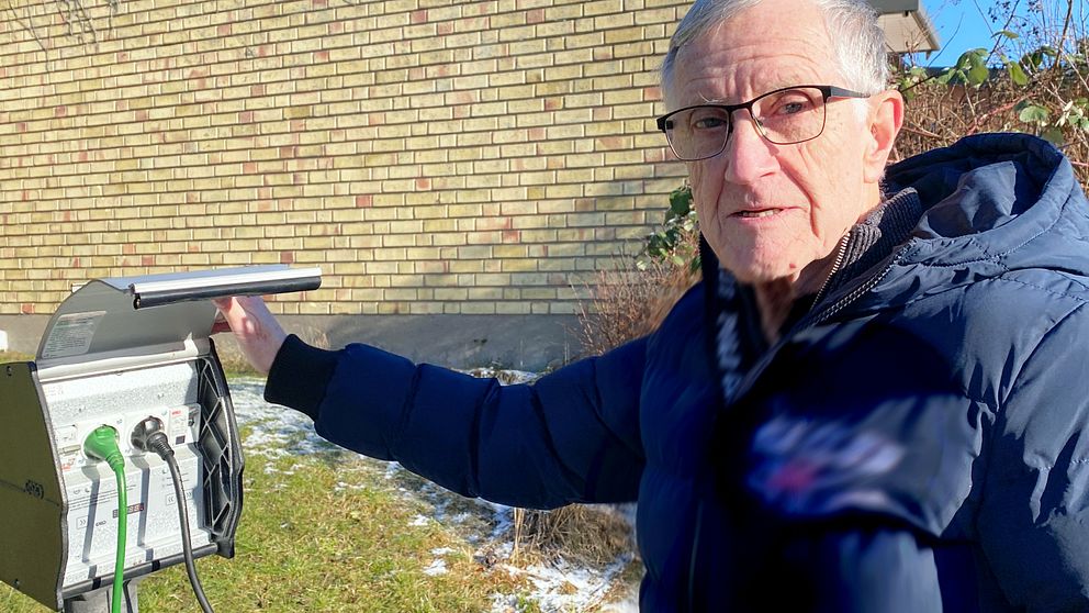 Person i vinterjacka pekar på elbilsladdare bredvid tegelbyggnad. Snö på marken.